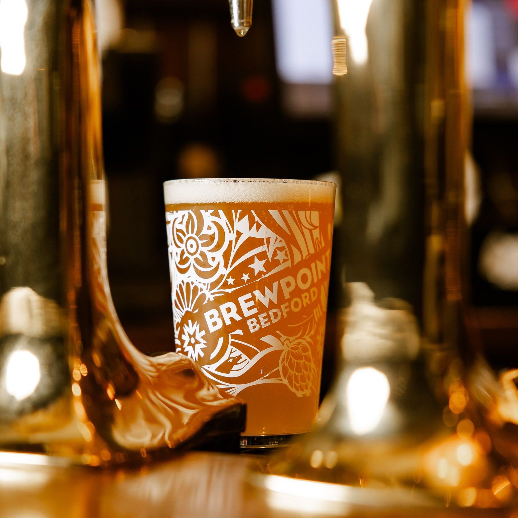 A picture of a Brewpoint beer next to a beer font on the bar. The glass is decorative dawning the logo of Brewpoint Bedford.