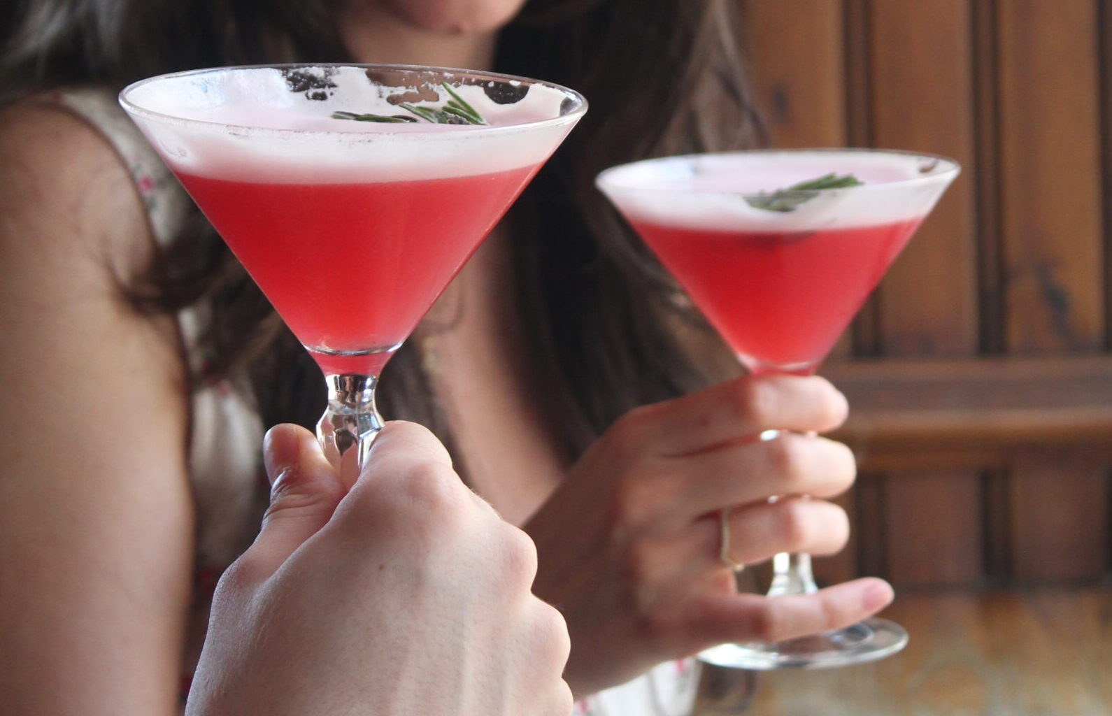 2 Cocktails on a 2 for 16.5 euro offer being held up. The cocktails are a pale red colour with a rosemary garnish.